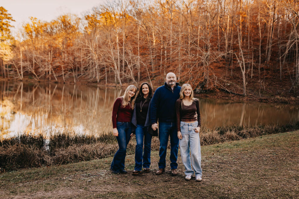 Family with teens smiling together in front of pond with fall foilage in backdrop