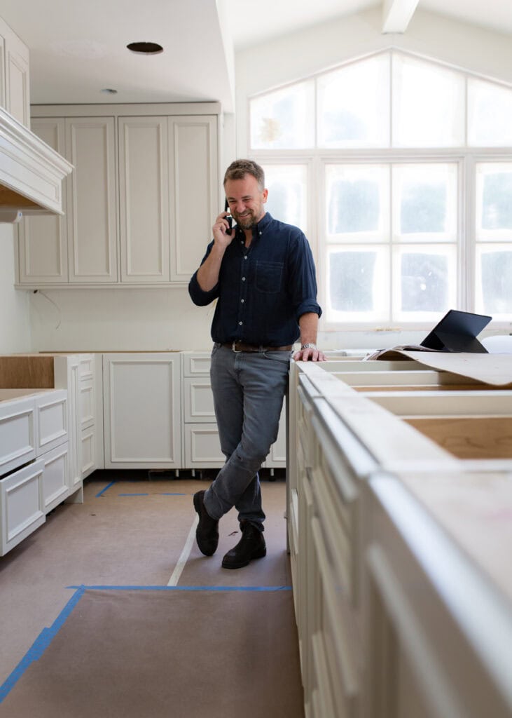 Man on phone in a bright, modern kitchen with large windows and white cabinetry.